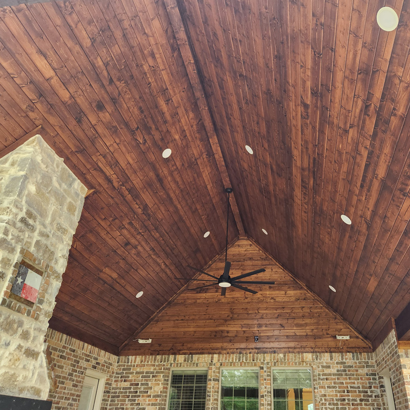 A vaulted, tongue and groove wood ceiling with a warm brown stain is the centerpiece of this outdoor living space. Numerous recessed lights are installed throughout the ceiling, providing ample illumination. A black, six-bladed ceiling fan hangs from the peak of the roof. The walls are a mix of brick and stone. This image demonstrates the high-quality lighting and electrical work provided by Eiland Electric.