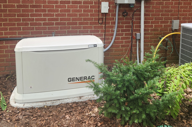A beige Generac home backup generator is installed on a concrete pad next to a red brick house. A gray metal electrical panel and a utility meter are mounted on the wall above and to the right of the generator, with conduit and wiring running between them and the generator. To the right of the generator is a large air conditioning unit. The ground is covered with mulch and various plants are visible around the equipment.