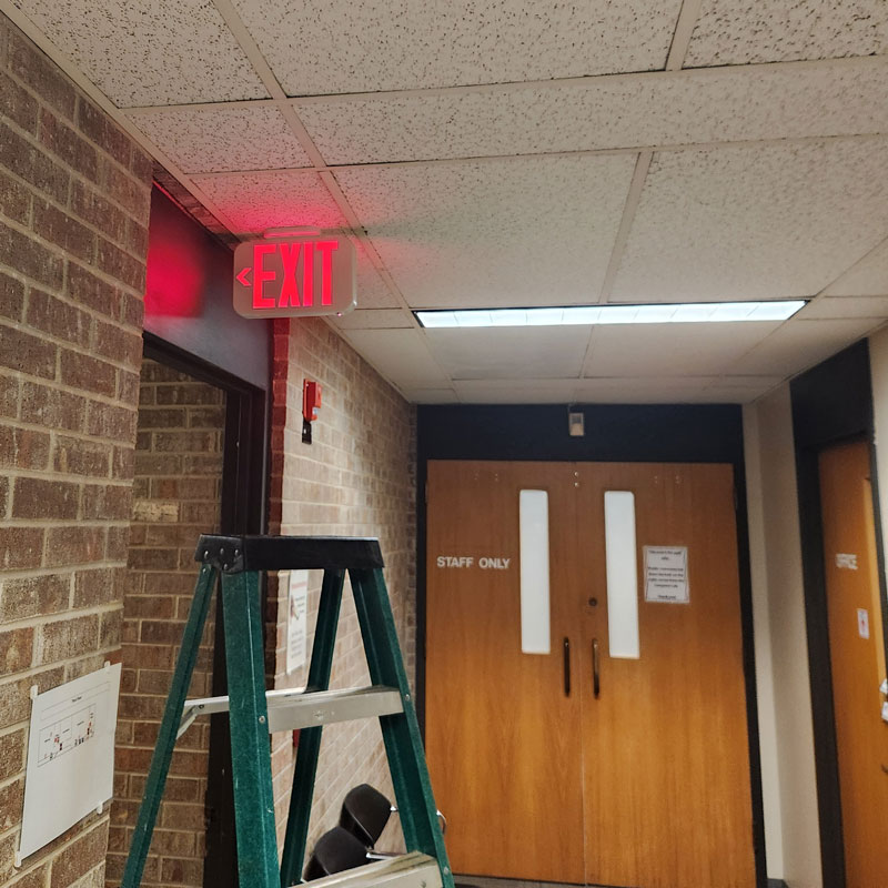 A hallway in what appears to be a commercial or institutional building. The hallway has a drop ceiling with fluorescent lighting. A red illuminated "EXIT" sign is installed on the ceiling near a corner, directing people out. A brick wall lines the left side of the hallway, and a set of double wooden doors with a "STAFF ONLY" sign is visible in the background. A green stepladder is positioned beneath the exit sign, suggesting Eiland Electric's work on the lighting installation.